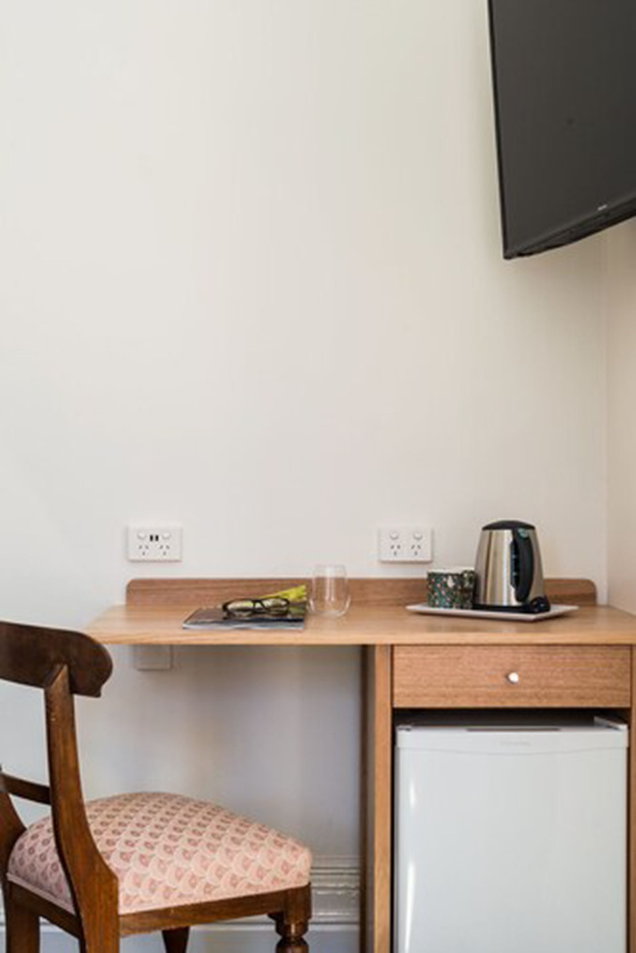 A compact room with a wooden desk, a small refrigerator, a chair with a patterned cushion, a wall-mounted TV, and a kettle on the desk.