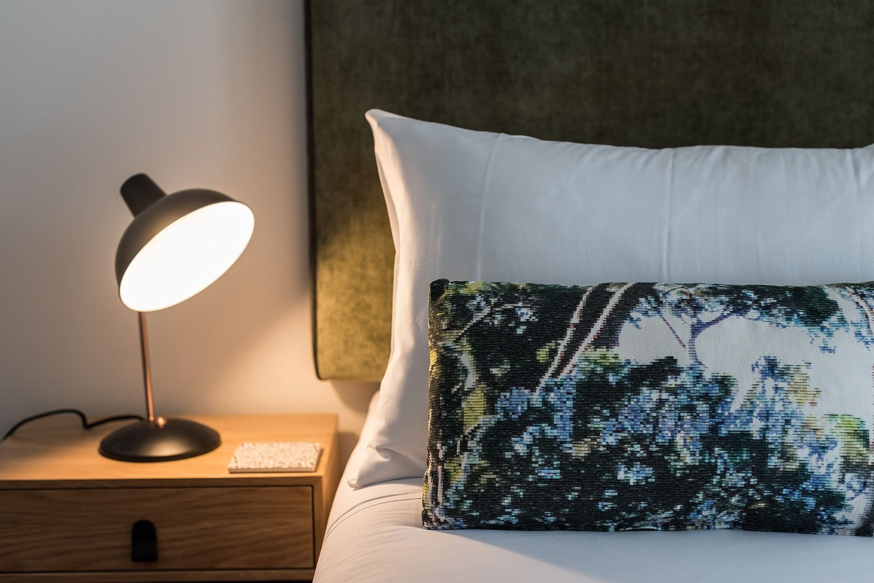 A neatly made bed with a white pillow and a patterned pillow, a bedside lamp, and a wooden nightstand with a keyboard.