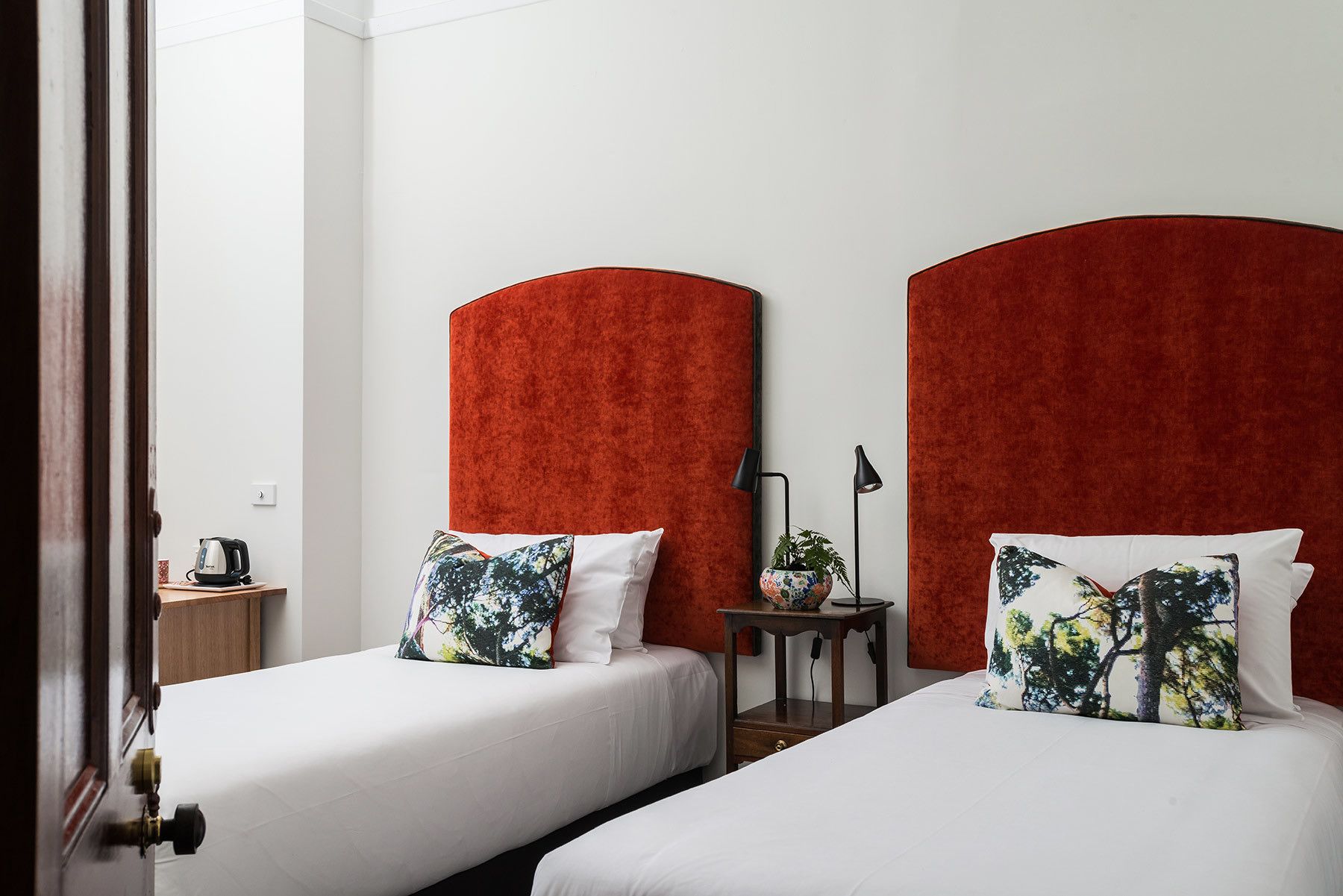 Habitación de hotel con dos camas, cada una con cabecero rojo y ropa de cama blanca. Se observa una mesita de noche con una lámpara y una planta decorativa.