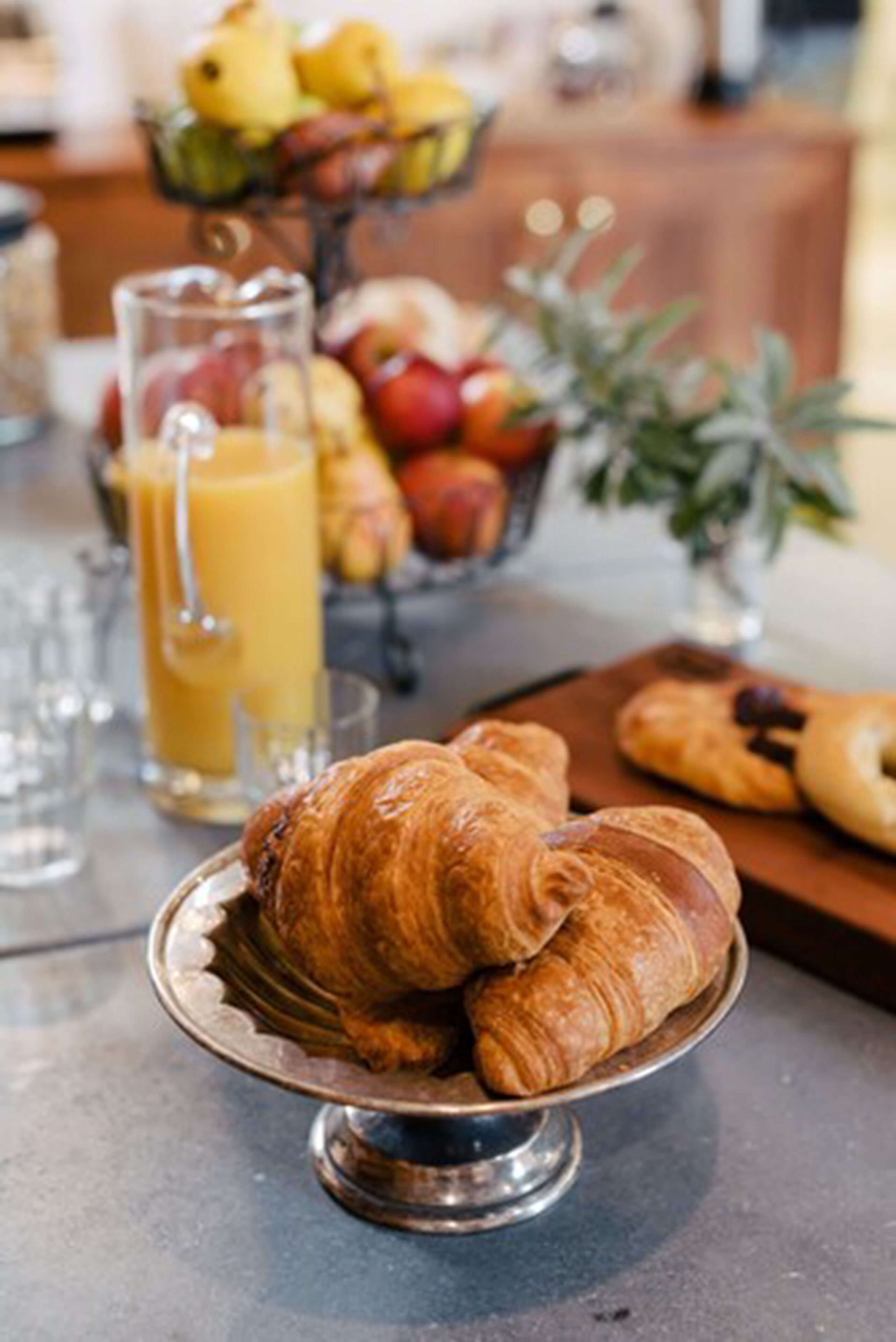 A breakfast setting with croissants on a metal stand, a fruit bowl, and a glass of orange juice.