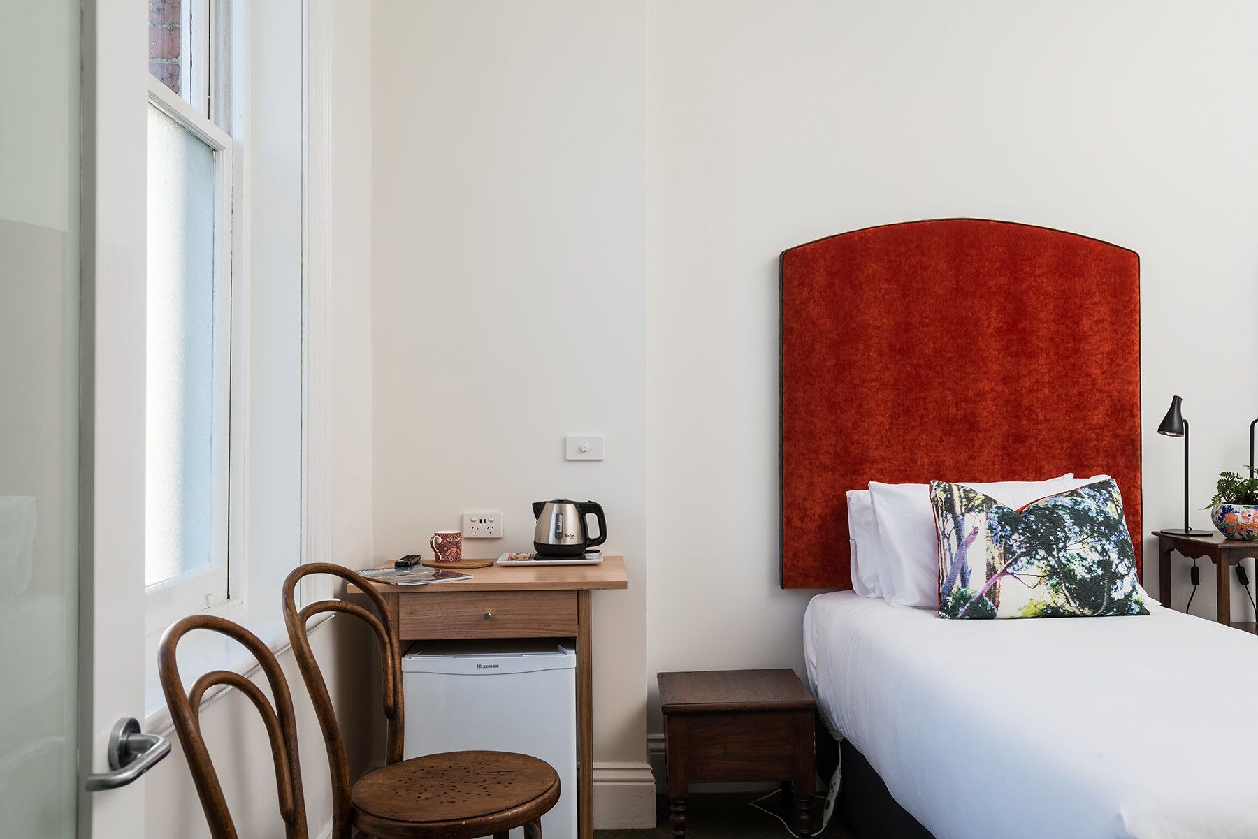 A hotel room with a bed, a desk with a kettle, a chair, and a small bedside table.