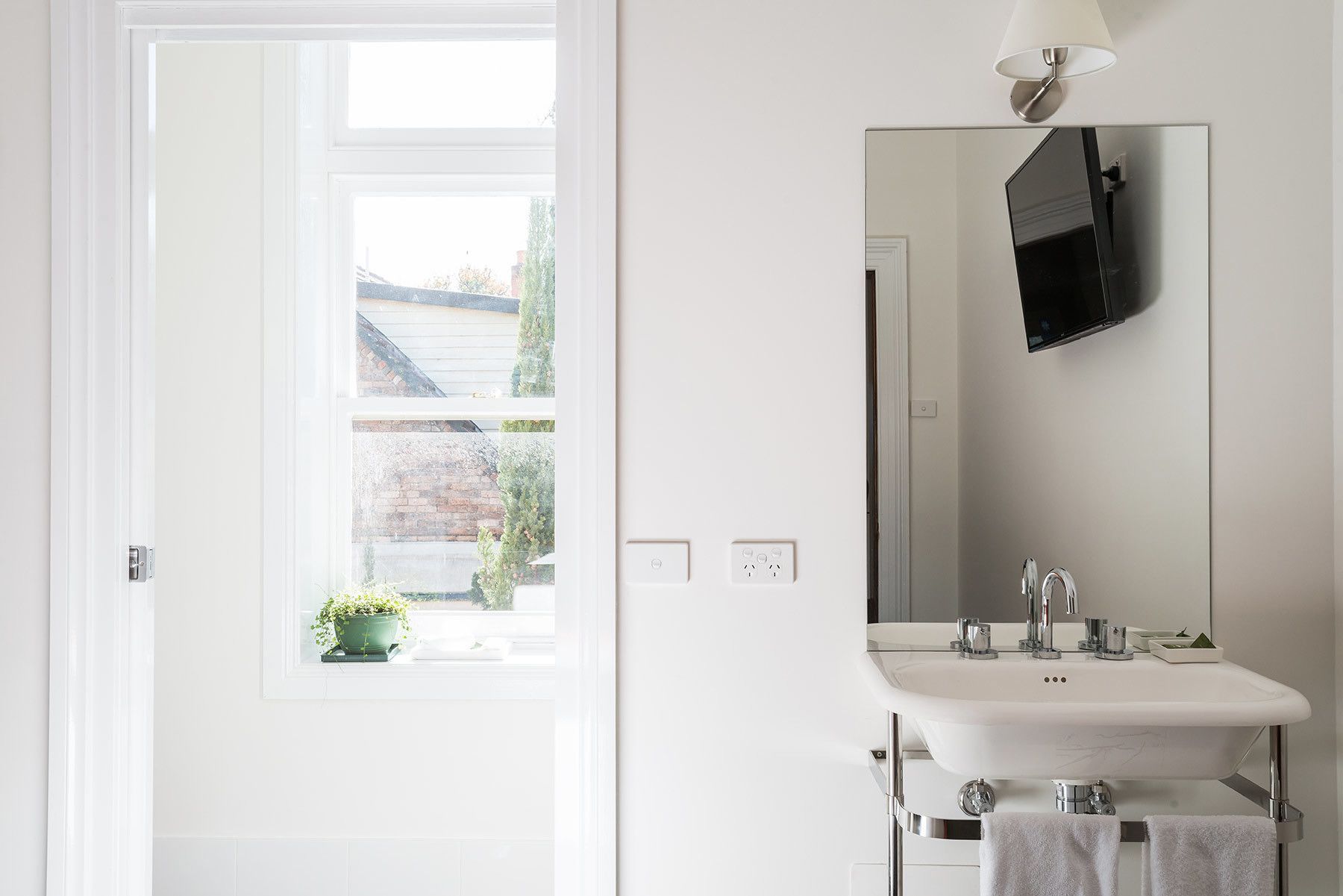 Un baño con un lavamanos blanco, un espejo, un televisor montado en la pared y una ventana que ofrece vistas a una casa y vegetación.