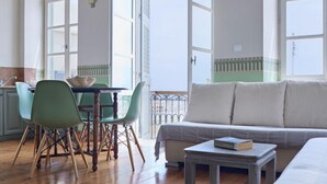 A spacious room with a wooden floor, a dining table with chairs, a sofa, and large windows providing natural light. - Guesthouse Lila (Syros)