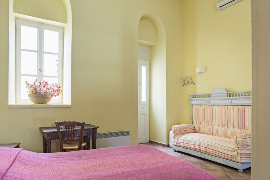 A room with a bed, a desk, a chair, a sofa, and a window with a flower arrangement.