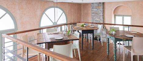 A dining area with wooden tables and white chairs, a stone wall, and large arched windows.