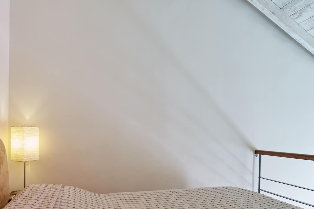 Quarto com cama, mesa de cabeceira, abajur e teto inclinado.