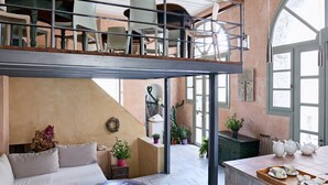 A spacious loft-style living area with a high ceiling, a mezzanine level, and a large window providing natural light. - Guesthouse Lila (Syros)