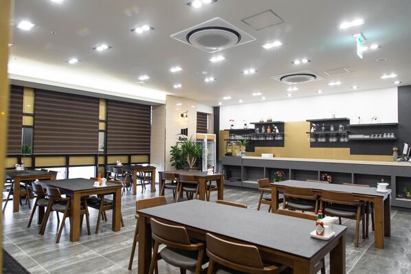 Un restaurante moderno con mesas y sillas de madera, una barra con electrodomésticos de cocina y un amplio ventanal con persianas.