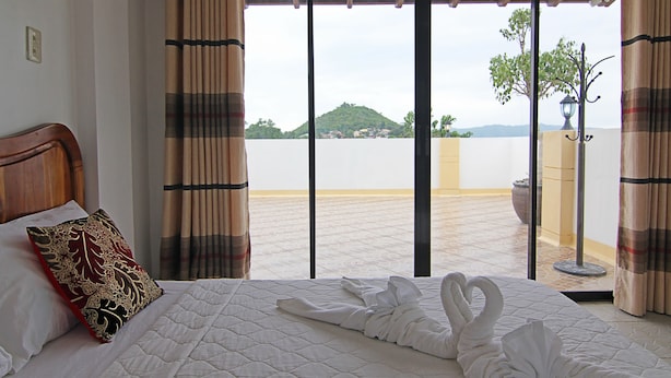 A hotel room with a bed, curtains, and a view of a cityscape through large windows. - Darcey Residences (Tagbilaran)