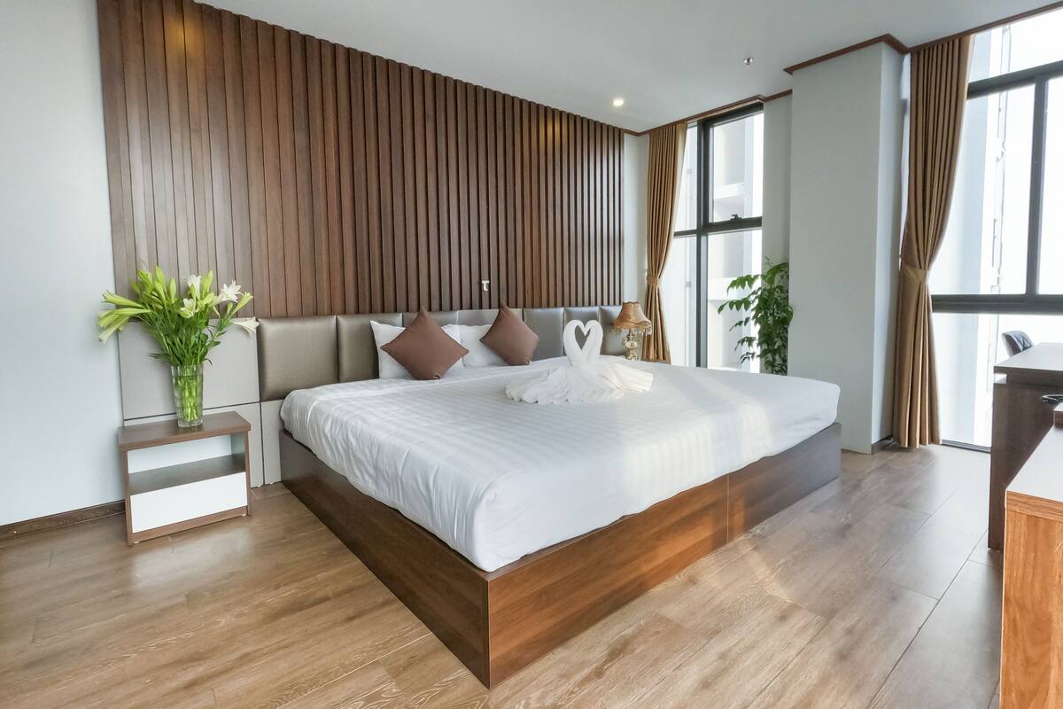 A modern bedroom with a large bed, wooden panel wall, and a small bedside table with a vase of flowers.