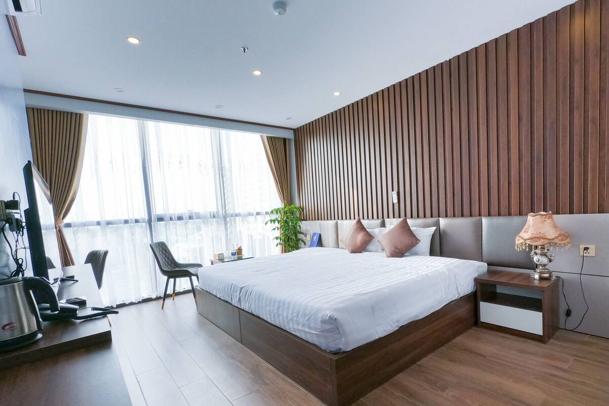 A modern hotel room with a large bed, a desk with a chair, a TV, and a large window with curtains.