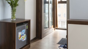 A hotel room with a wooden floor, a refrigerator with a children's meal sticker, a vase with flowers, and a view of a building through a glass door. - Hello Hotel (Vo Cuong)