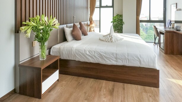 A modern bedroom with a large bed, wooden paneling on the wall, a bedside table with a vase of flowers, and a desk area with a chair. - Hello Hotel (Vo Cuong)