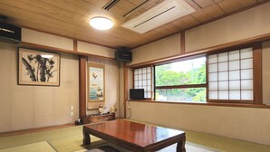 Habitació japonesa tradicional amb sòls de tatami, una taula de fusta baixa, un coixí de tatami, una obra d'art emmarcada, una televisió i una finestra amb vista a vegetació.