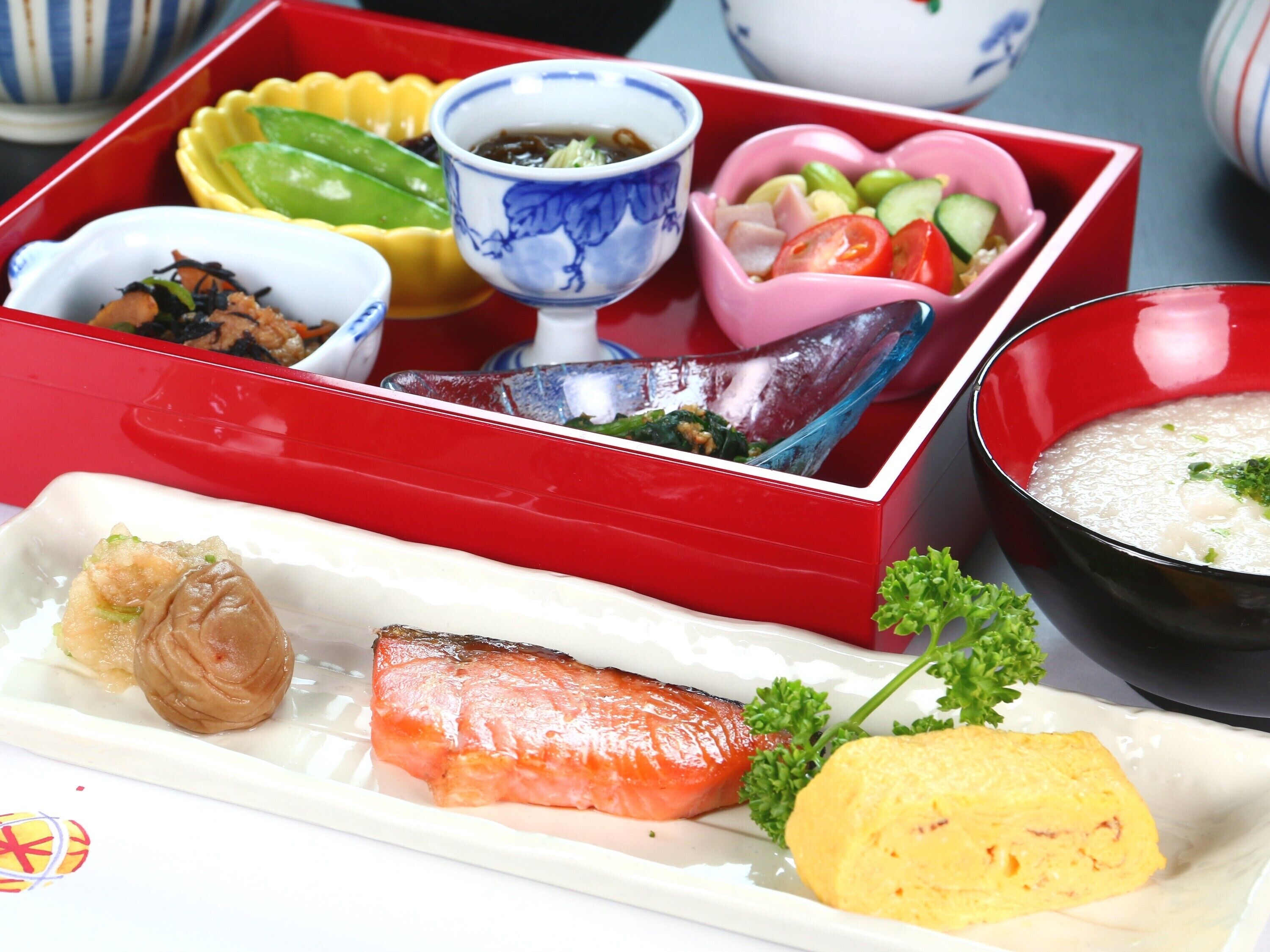 Una comida tradicional japonesa con varios platos en una caja lacada de color rojo, incluyendo salmón, verduras encurtidas y un tazón de sopa.