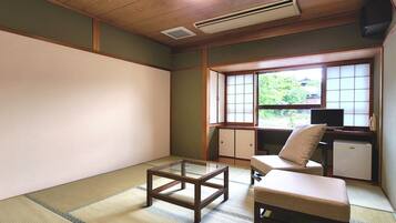 Um quarto japonês tradicional com piso de tatame, uma mesa baixa, uma cadeira estofada, uma escrivaninha com um computador e uma janela com vista para o verde.