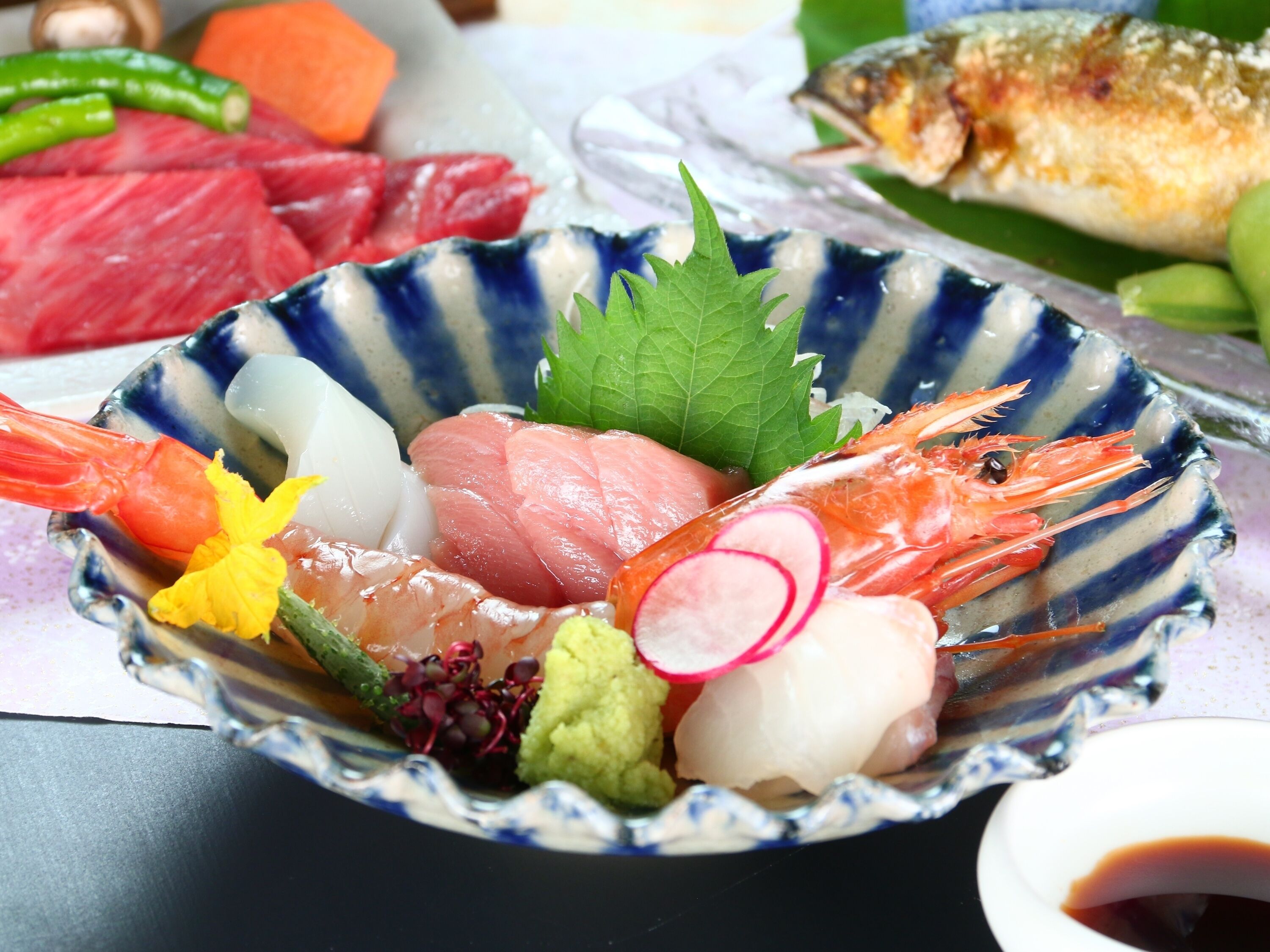 Eine Schüssel mit verschiedenen Sashimi und Meeresfrüchten, darunter Garnelen, Fisch und Masaito, garniert mit Gemüse und Kräutern.