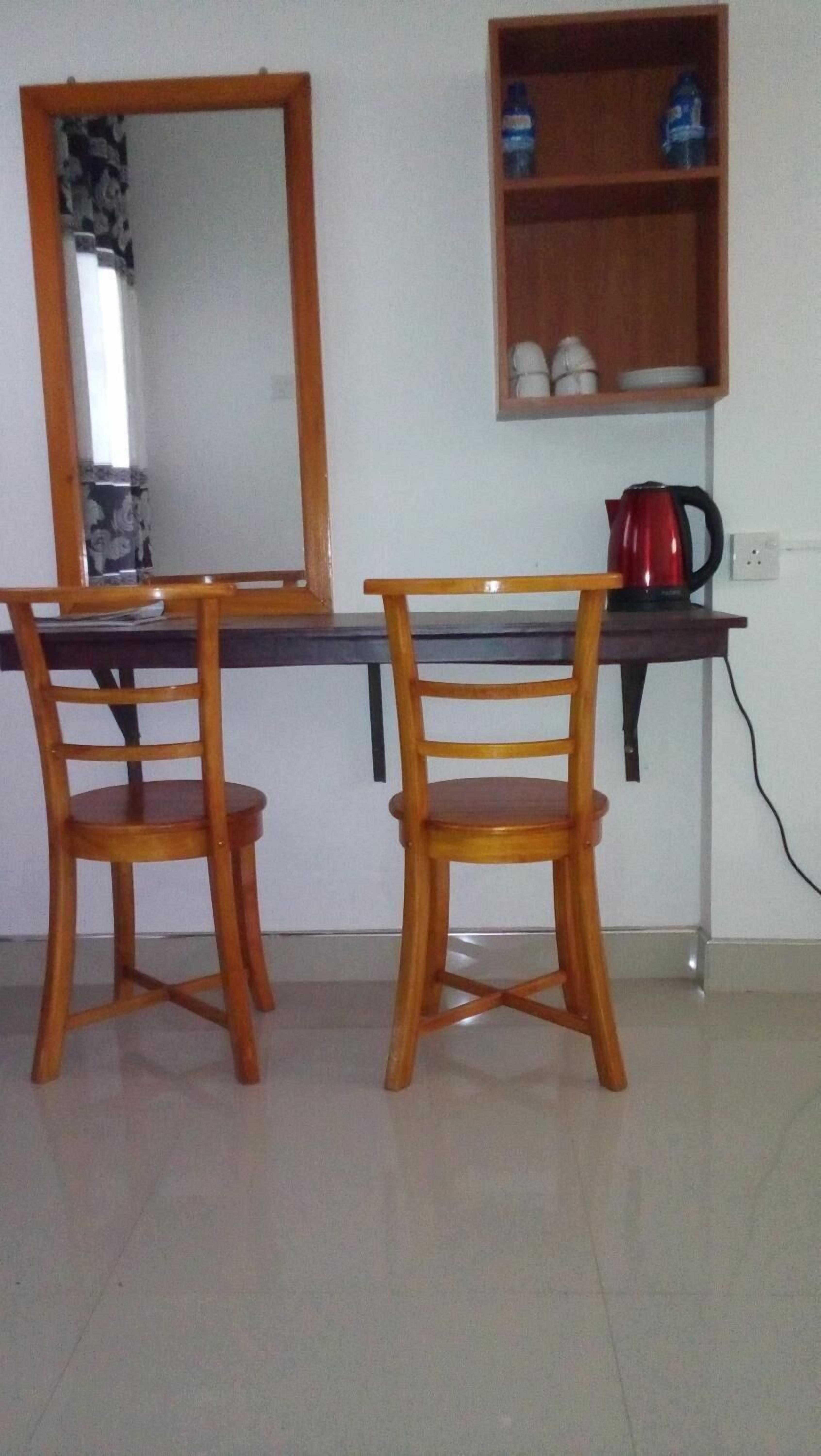 Une pièce comprenant deux chaises en bois, un bureau, une bouilloire rouge et une étagère avec des bouteilles d’eau et des tasses.