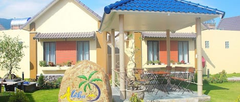 A hotel with a covered outdoor seating area, a stone sign with the hotel name, and a well-maintained garden.