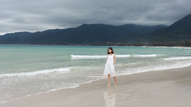 Une femme vêtue d’une robe blanche se tient sur une plage, avec une eau turquoise et des montagnes en arrière-plan.