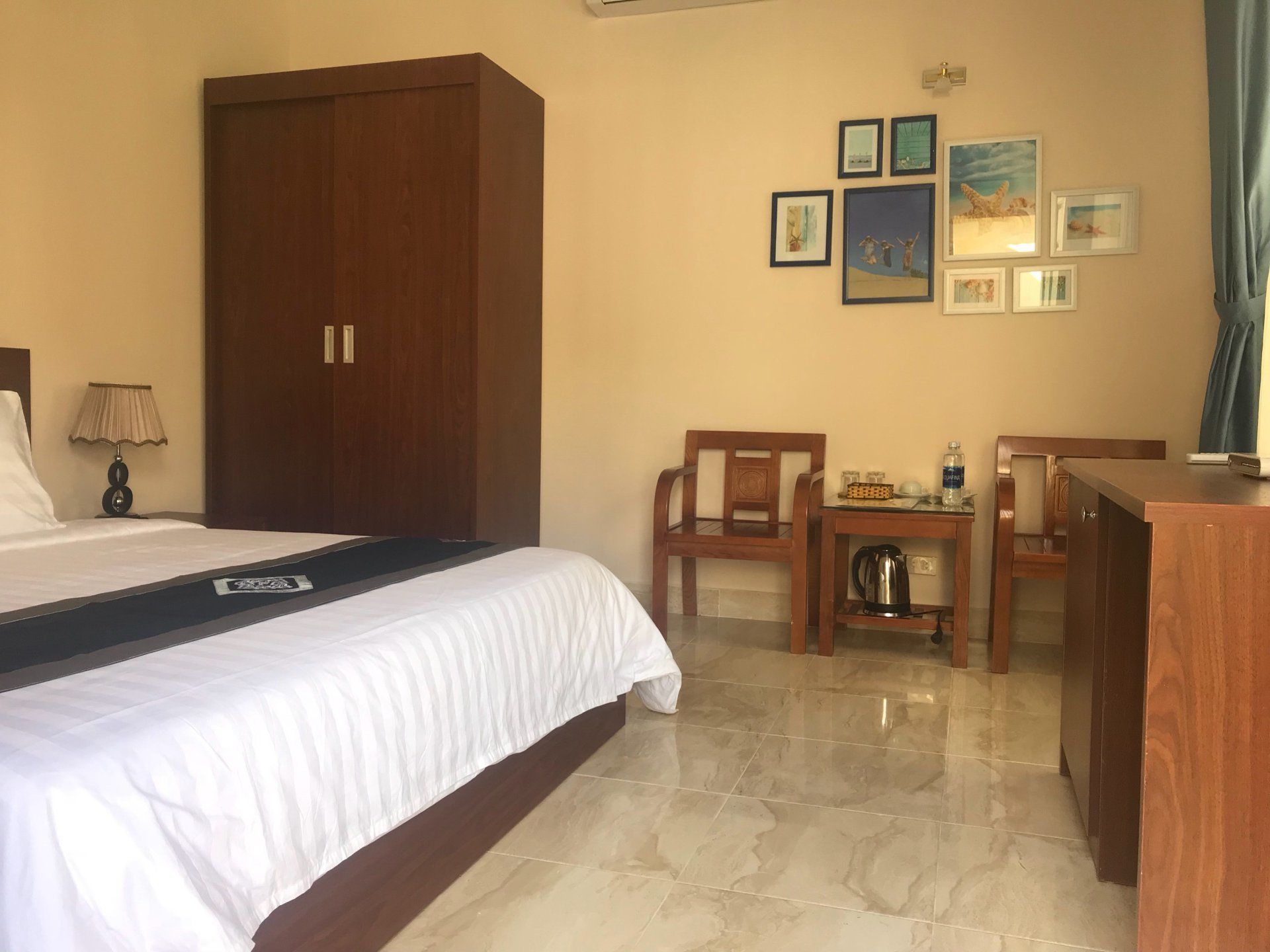 A hotel room with a bed, a wooden wardrobe, a desk with a chair, a coffee maker, and framed pictures on the wall.