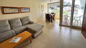Ein Wohnzimmer mit grauem Sofa, einem Holztisch und einem Essbereich mit Tisch und Stühlen. An der Wand hängen zwei gerahmte Kunstwerke, und eine Glastür führt auf einen Balkon mit weißen Plastikstühlen.