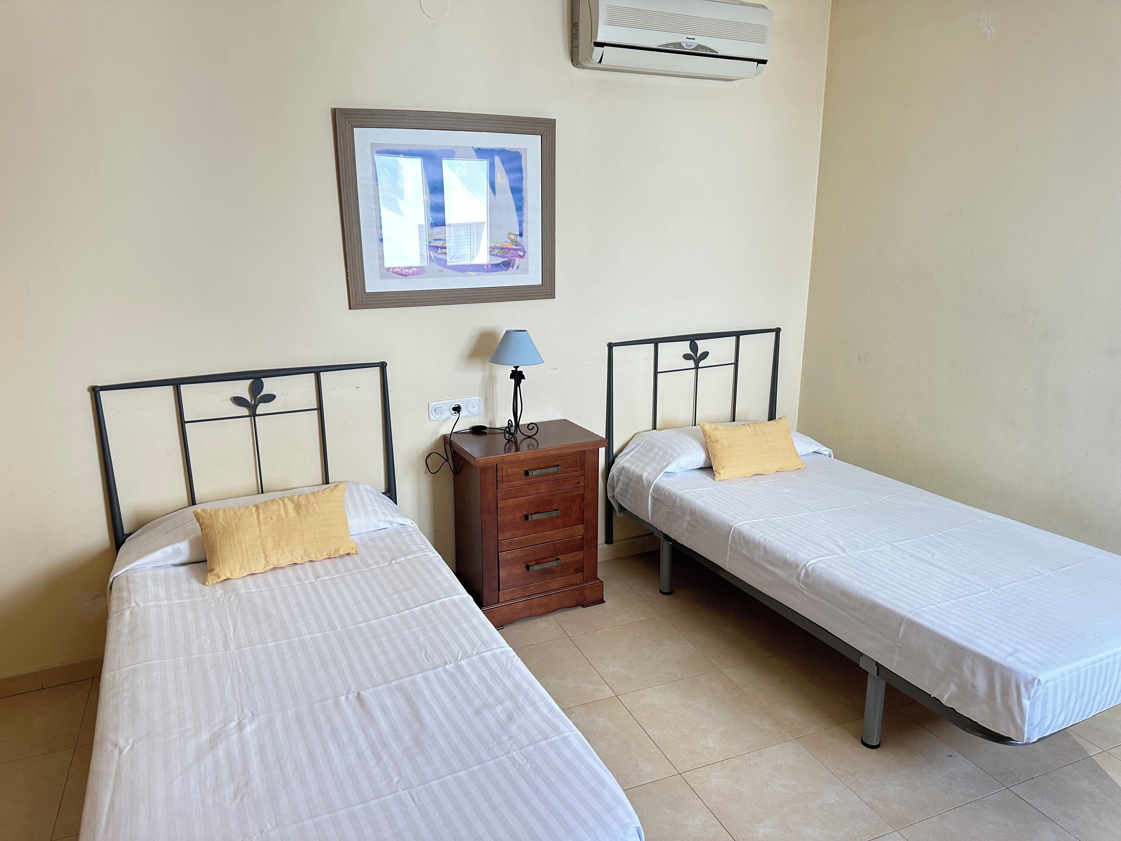 Un dormitorio con dos camas individuales, una mesita de noche con lámpara, un cuadro en la pared y un aire acondicionado instalado en el techo.