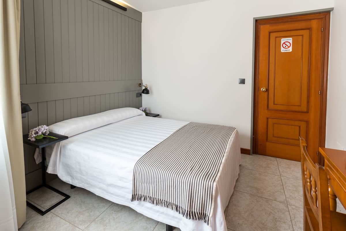 Un lit bien fait, recouvert d’une couverture à rayures, une table de chevet avec une lampe et une porte en bois sur laquelle est apposé un panneau « interdiction de fumer ».