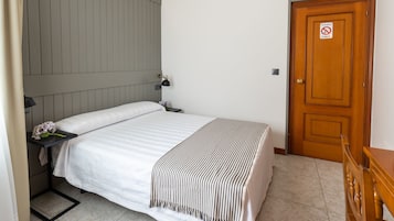 Una cama bien hecha con una manta a rayas, una mesita de noche con una lámpara y una puerta de madera con un cartel de 'no fumar'.
