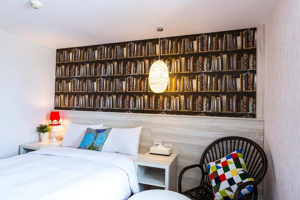 Quarto de hotel com uma grande estante de livros, uma cama com roupa de cama branca, um abajur vermelho e uma cadeira preta com almofada colorida.