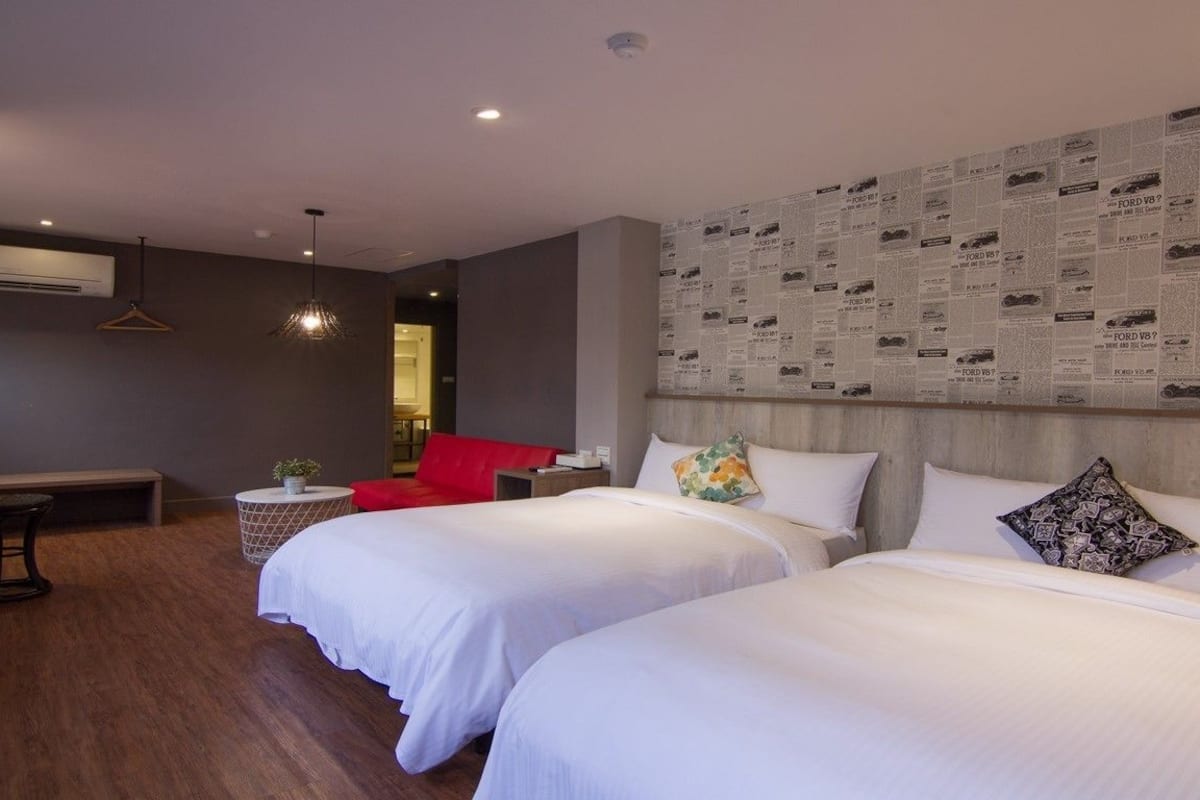 Quarto de hotel com duas camas, uma cadeira vermelha, uma mesa pequena e uma parede decorada com recortes de jornal.
