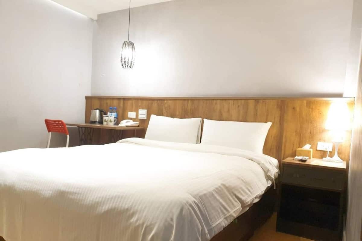 Quarto de hotel com uma cama grande, cabeceira de madeira, escrivaninha com cadeira, mesa de cabeceira com abajur e luminária pendente.