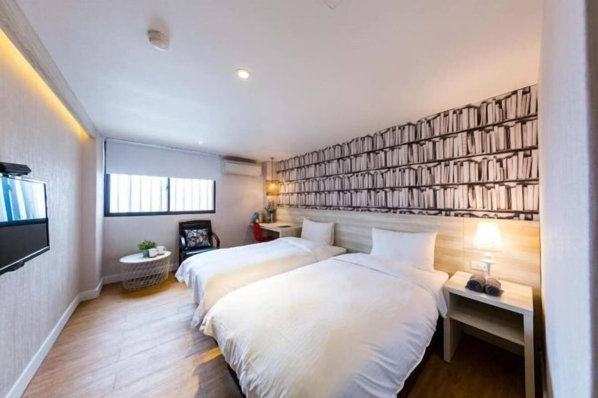 Quarto de hotel com duas camas, uma estante de livros, uma TV e uma pequena mesa redonda.