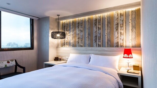 A modern hotel room with a large bed, a nightstand, a red lamp, and a view of the city through a large window. - Artch Inn (Luodong)