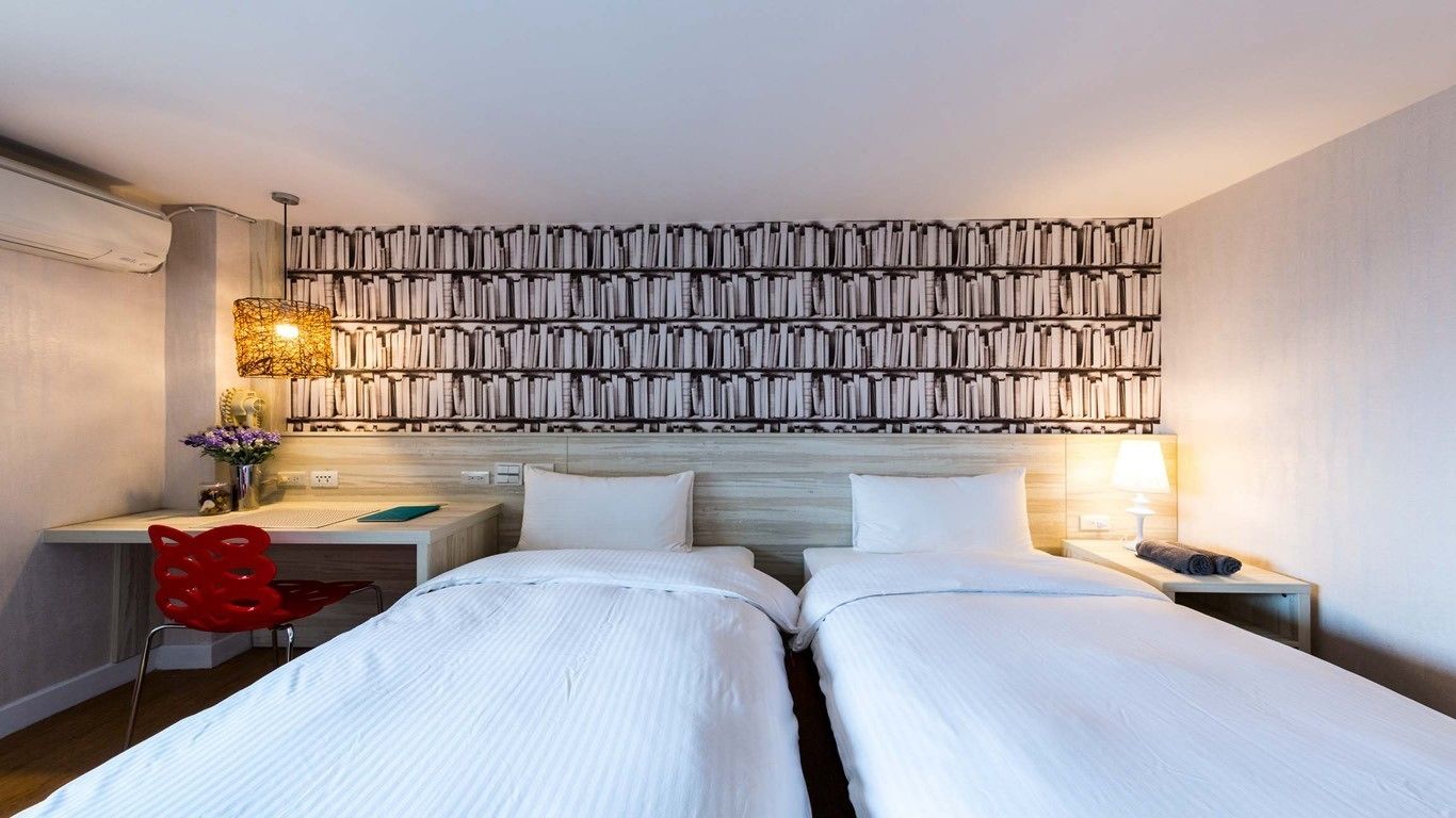 A hotel room with two beds, a desk, a red chair, and a wall-mounted bookshelf.