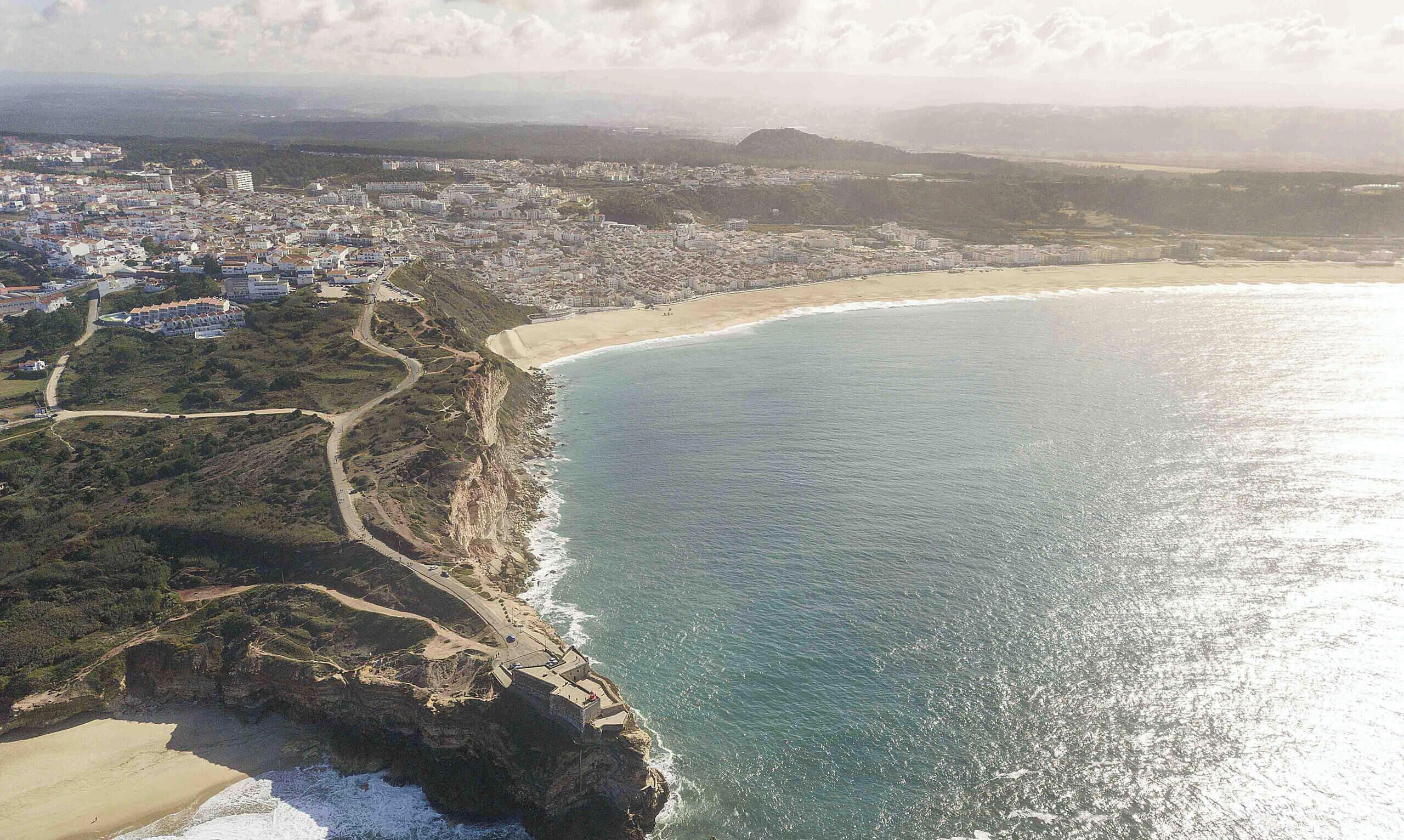 Vista aerea di una cittadina costiera con spiaggia, una strada tortuosa e un sentiero a picco sulla scogliera.