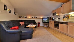A cozy living room with a black leather sofa, a small coffee table, and a TV. There is a kitchen area with wooden cabinets and a microwave.