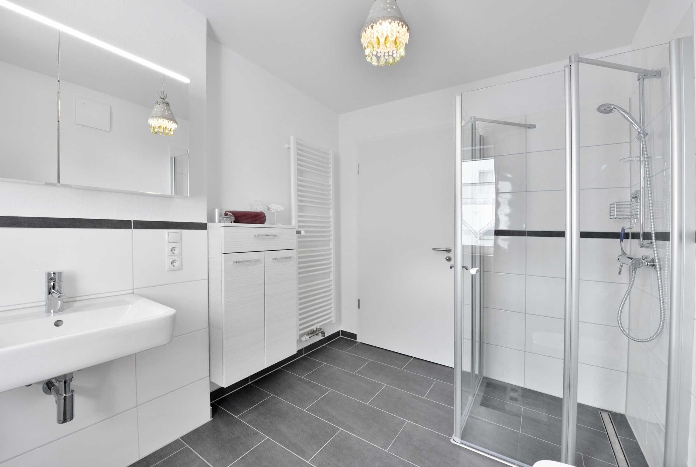 A modern bathroom with a glass shower enclosure, a white sink, and a chandelier.