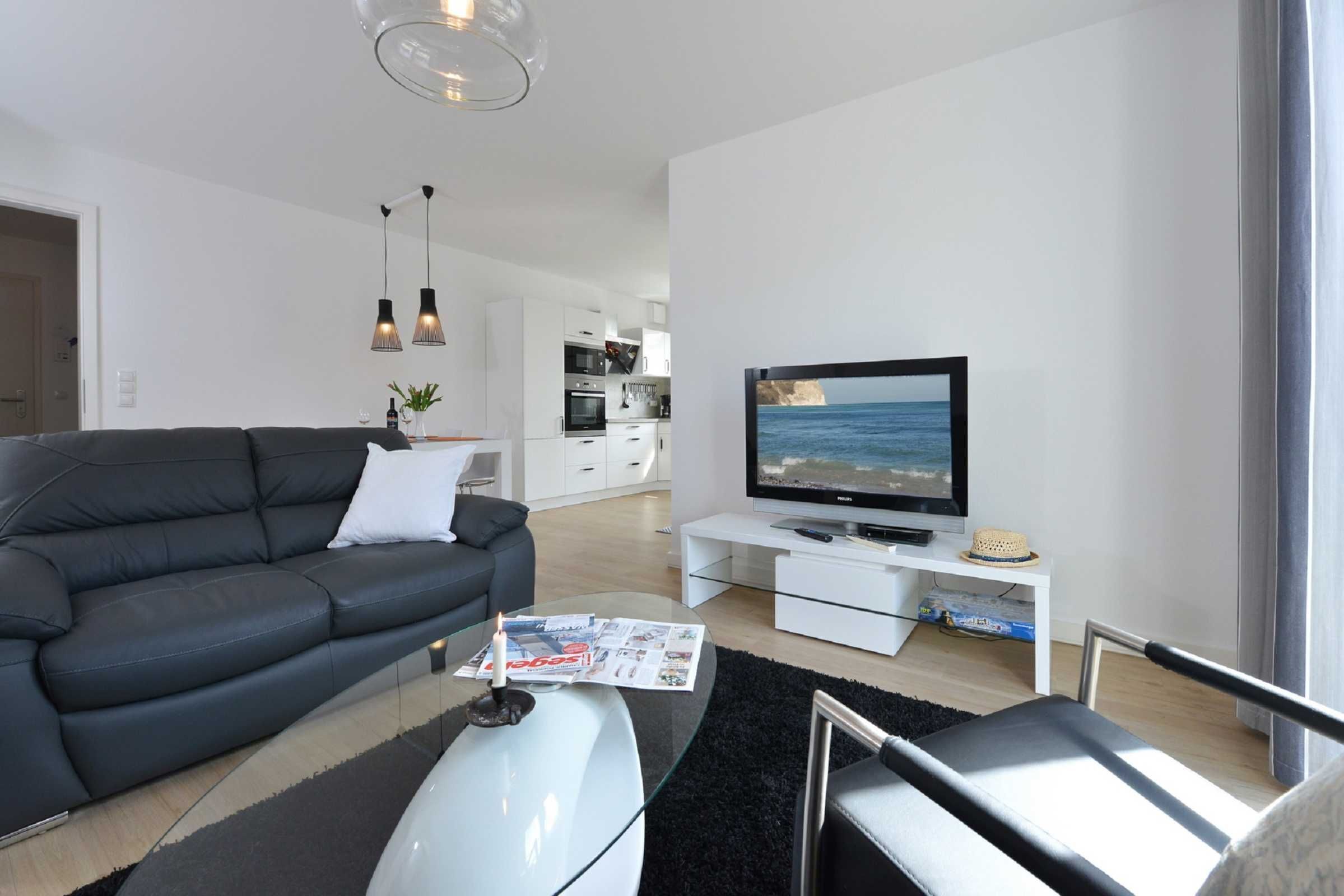 Una sala moderna con un sofá de cuero negro, una mesa de centro de cristal y un televisor de pantalla plana sobre un mueble blanco.