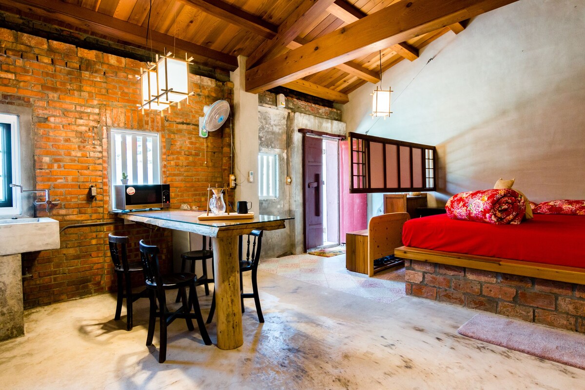 A rustic room with a brick wall, wooden ceiling beams, a dining table with chairs, a bed with a red bedspread, and a small wooden bench.