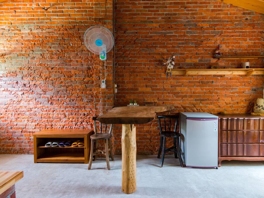 A rustic room with exposed brick walls, a wooden table, two chairs, a small wooden shelf with shoes, a refrigerator, and a fan mounted on the wall.
