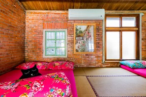 Ein Zimmer mit Backsteinwand, einem Fenster mit Jalousien, einem Bett mit blumigem Bettzeug und einer schwarzen Katze auf dem Bett, sowie einem kleinen Fenster mit Blick auf Hühner.
