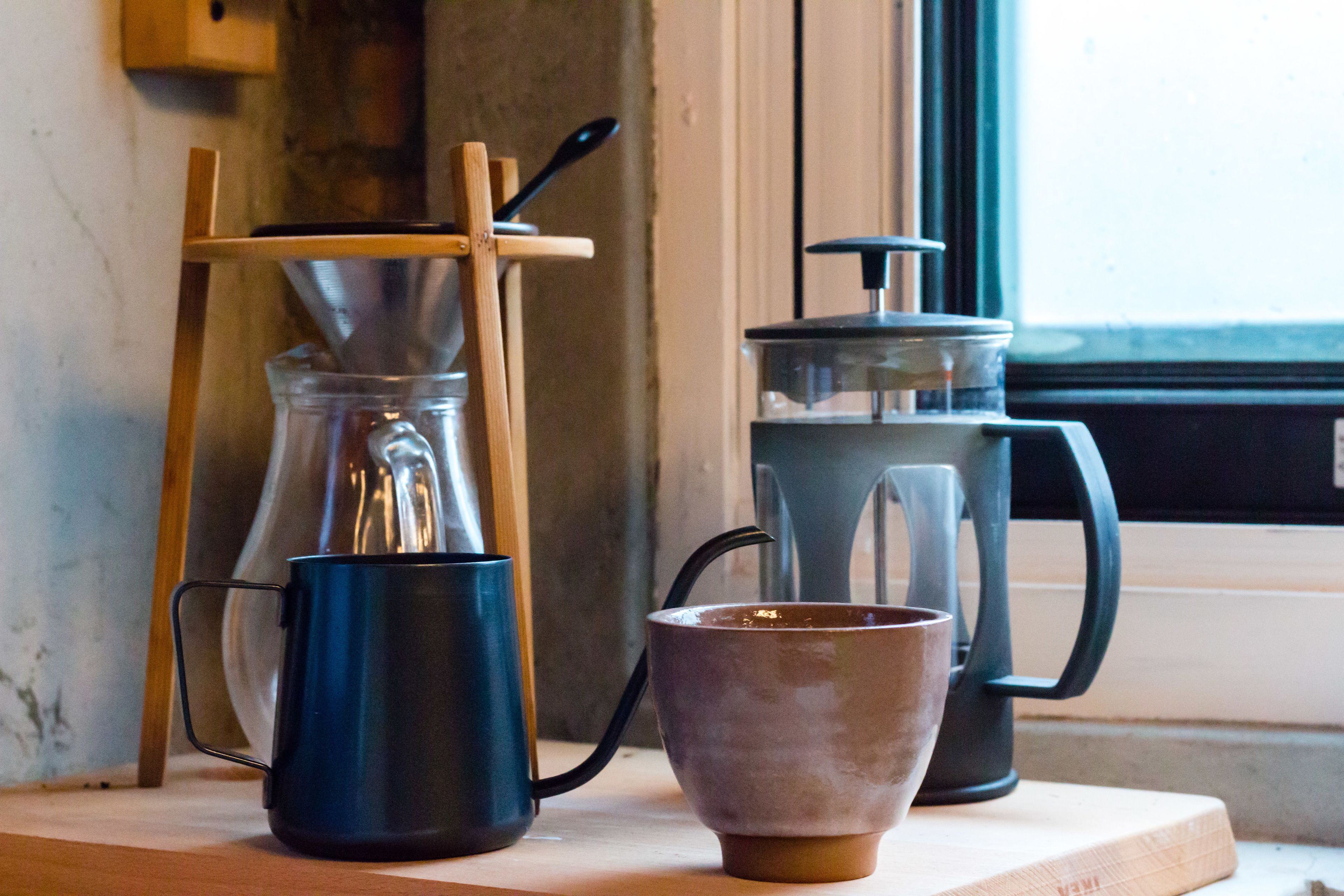 Una cafetera, una taza y una jarra de vidrio sobre una mesa de madera cerca de una ventana.