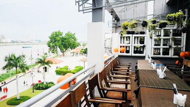 A balcony with wooden chairs and tables, potted plants, and a view of a river and cityscape. - Tonle Khmer Guesthouse (Phnom Penh)