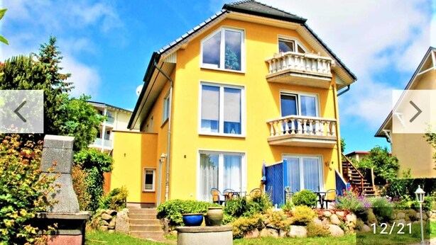 A two-story yellow building with a balcony on the second floor, surrounded by greenery and a stone structure.