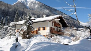 A chalet-style building with a sloped roof and wooden balconies, surrounded by snow-covered trees and a mountain in the background. - Vacation apartment Hoher Göll, 2-3 persons, 60 sqm, shower/WC, balcony, TV, W-LAN (Ramsau bei Berchtesgaden)