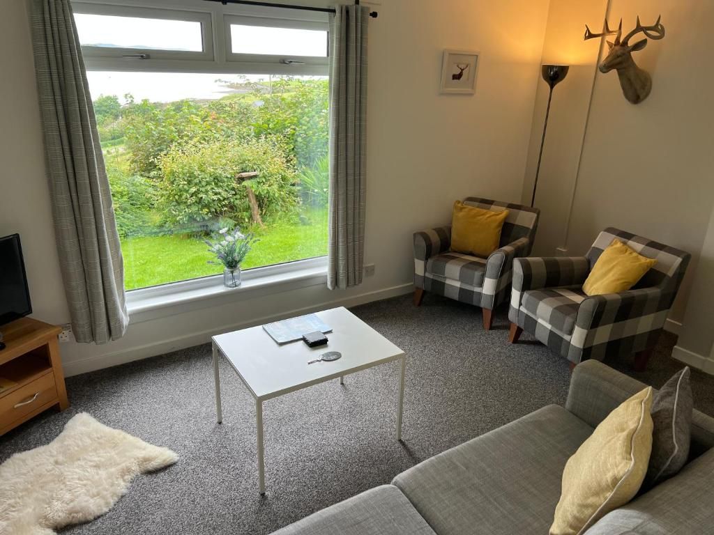 Un salon confortable avec un canapé, deux fauteuils, une table basse et un téléviseur. Une fenêtre donne sur un paysage verdoyant.