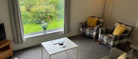 Un salon confortable avec un canapé, deux fauteuils, une table basse et un téléviseur. Une fenêtre donne sur un paysage verdoyant.