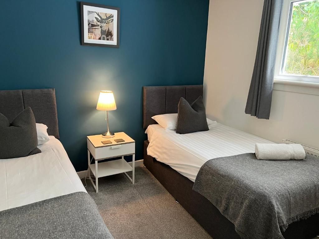 Quarto de hotel com duas camas, uma mesa de cabeceira com abajur, um quadro na parede e uma janela com cortinas.