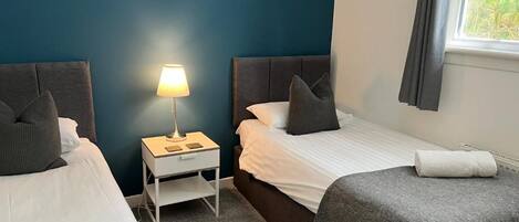 Quarto de hotel com duas camas, uma mesa de cabeceira com abajur, um quadro na parede e uma janela com cortinas.